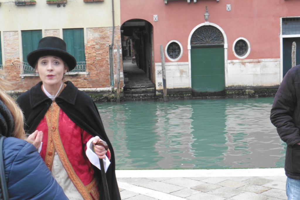 역사적인 복장을 한 사람이 오래된 건물을 배경으로 운하 옆에서 이야기를 나누고 있습니다. 다른 사람이 부분적으로 보입니다.