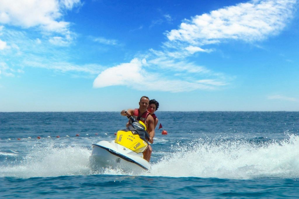 구름이 흩날리는 푸른 하늘 아래 탁 트인 물 위에서 노란 제트 스키를 타는 두 사람.