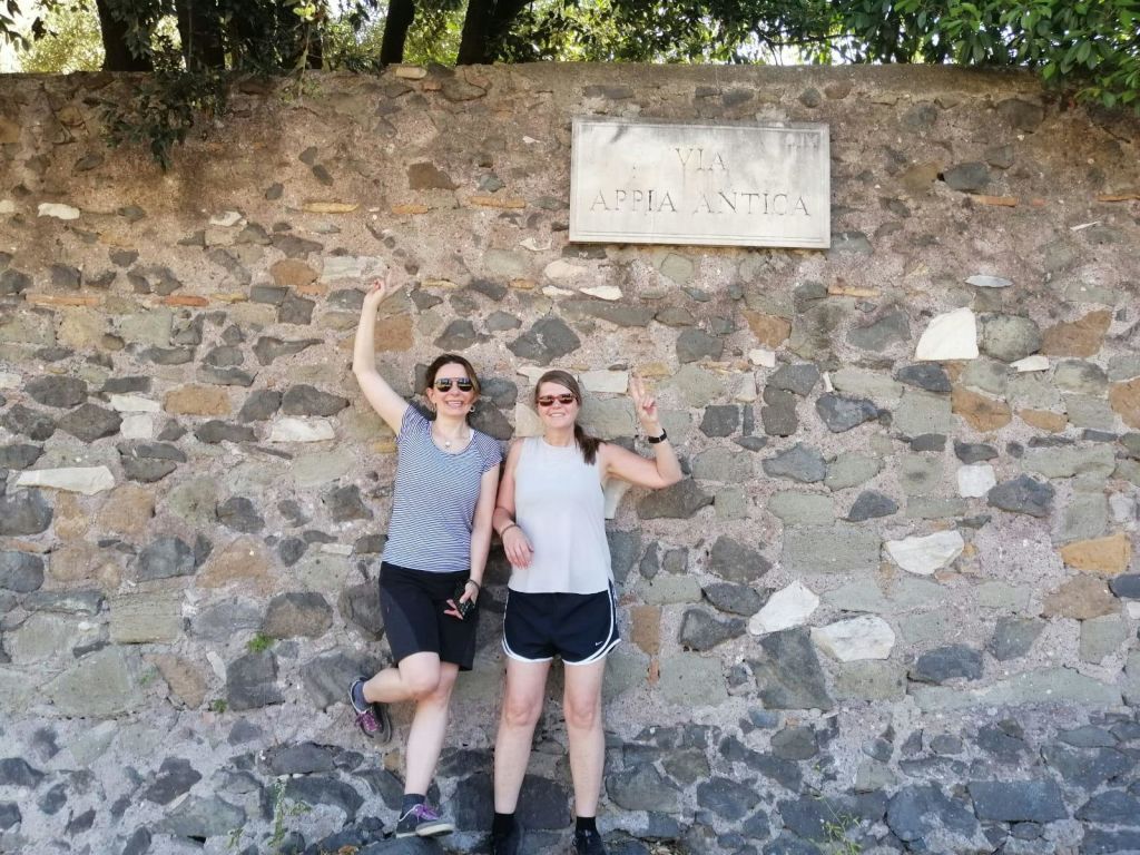 두 사람이 돌담 앞에 서서 "VIA APPIA ANTICA" 라고 적힌 표지판 아래에서 미소를 짓고 포즈를 취하고 있습니다.