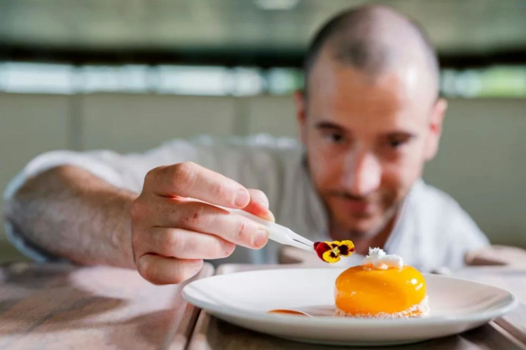 한 사람이 흰 포크로 노란색 유약 디저트 위에 조심스럽게 식용 꽃을 올려놓고 집중하고 있습니다.