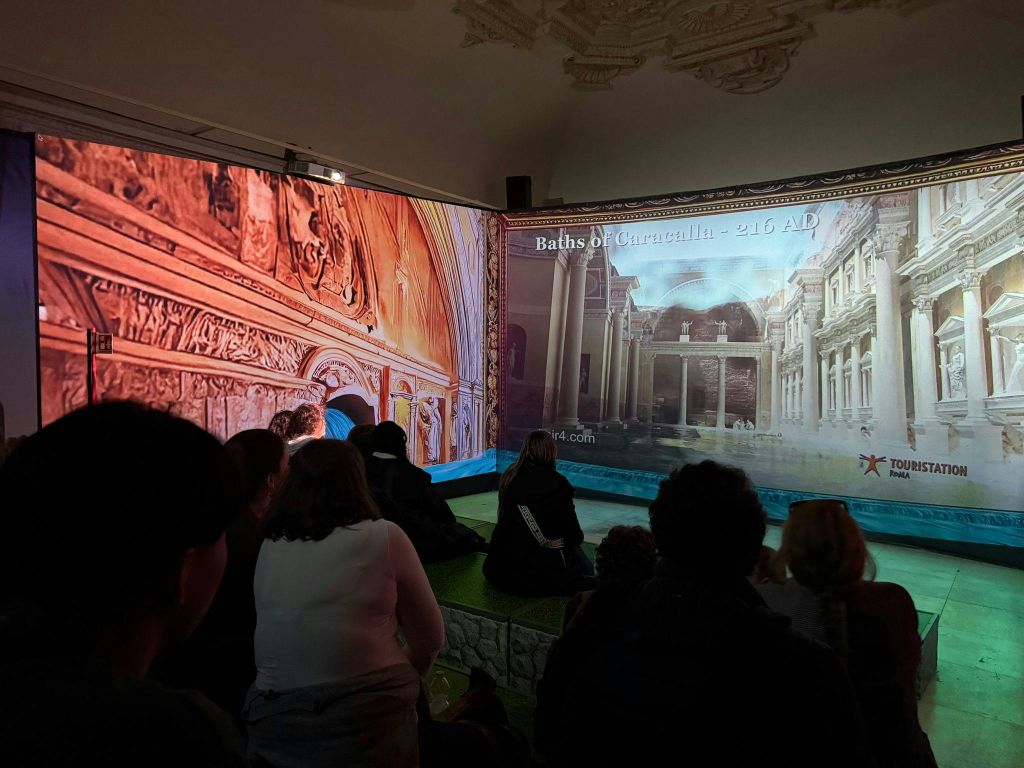 어두운 방에 앉아 '카라칼라의 목욕탕 - 서기 216년' 이라고 적힌 고대 로마 건축물을 보고 있는 한 무리의 사람들.