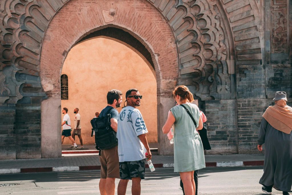 역사적인 아치형 관문 근처에 세 사람이 서서 물병을 들고 있습니다. 행인과 다른 아치가 배경에 보입니다.
