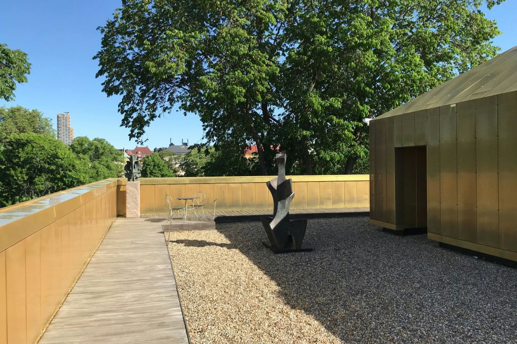 맑은 하늘 아래 나무와 황금 벽으로 둘러싸인 현대적인 조형물, 자갈, 작은 테이블 세트가 있는 옥상입니다.