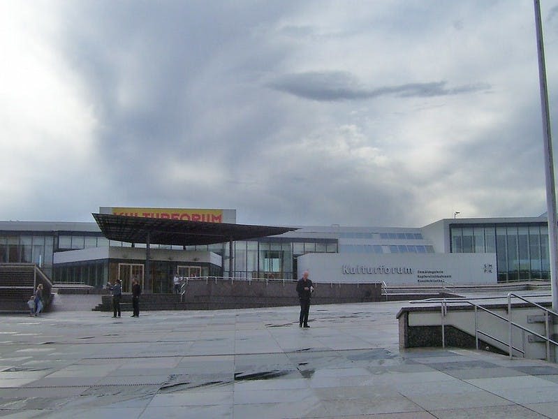 흐린 날 "Kulturforum" 이라고 적힌 표지판이 있는 현대적인 건물 앞의 넓은 광장에 서 있는 두 사람.