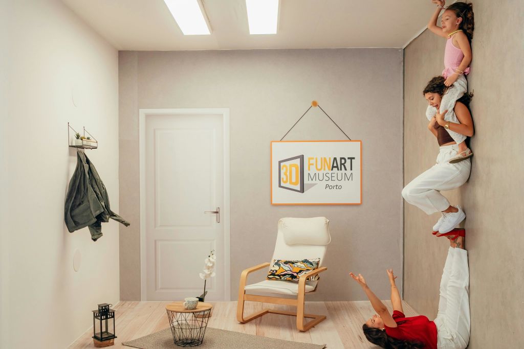 한 여성이 바닥에 누워 양손에 다른 여성을 안고 서 있습니다. 한 여성은 박물관 방에서 "3D FUN ART MUSEUM Porto" 간판이 있는 아이를 태우고 있습니다.