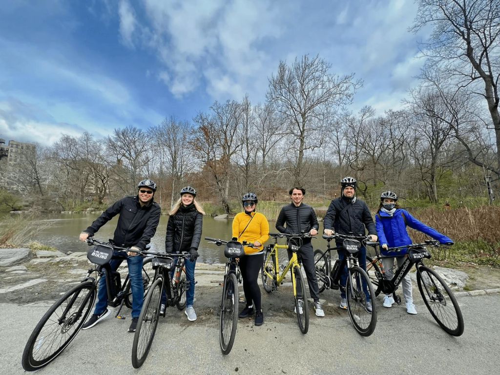 헬멧을 쓴 6명의 사람이 잎이 없는 나무 근처의 길에서 자전거를 타고 부분적으로 흐린 하늘 아래 작은 물줄기를 맞으며 포즈를 취하고 있습니다.