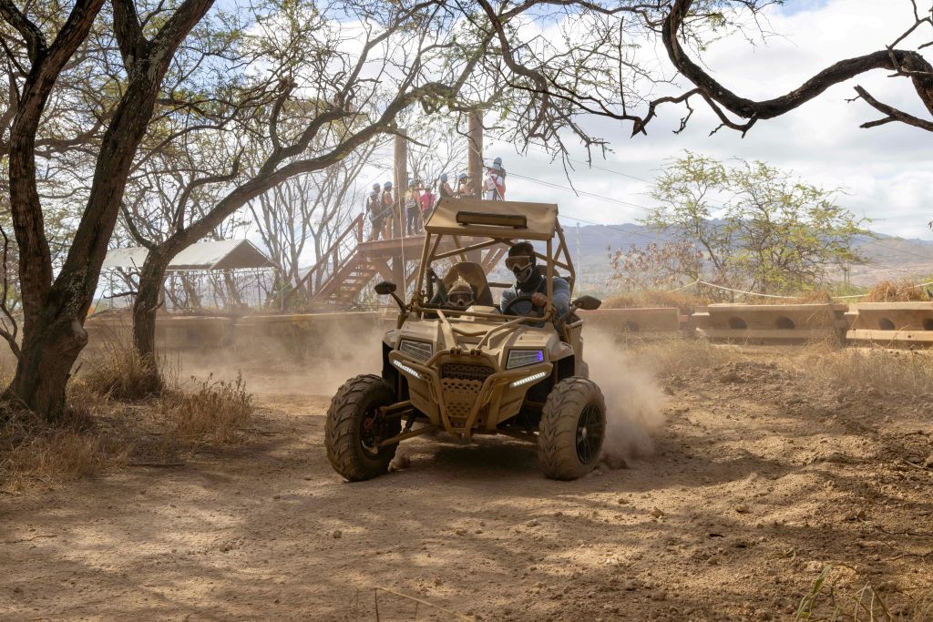 헬멧을 쓴 두 사람이 먼지가 쌓인 산책로를 따라 모래 언덕을 달리며 나무와 전망대를 배경으로 차를 몰고 있습니다.