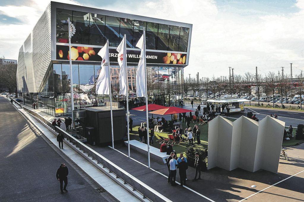 반사 유리와 깃발이 달린 현대적인 건물 근처의 좌석과 우산 아래에서 사람들이 어울리는 야외 장면.