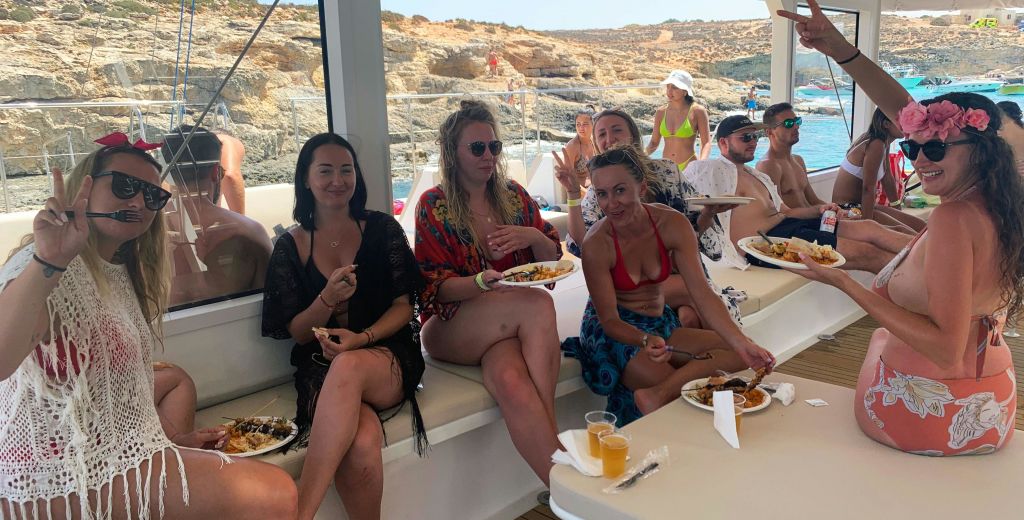 세 명의 여성이 보트에 앉아 음식을 즐기고 있습니다. 그들은 웃으며 캐주얼한 여름 복장을 하고 있습니다. 바위가 많은 해안선이 보입니다.