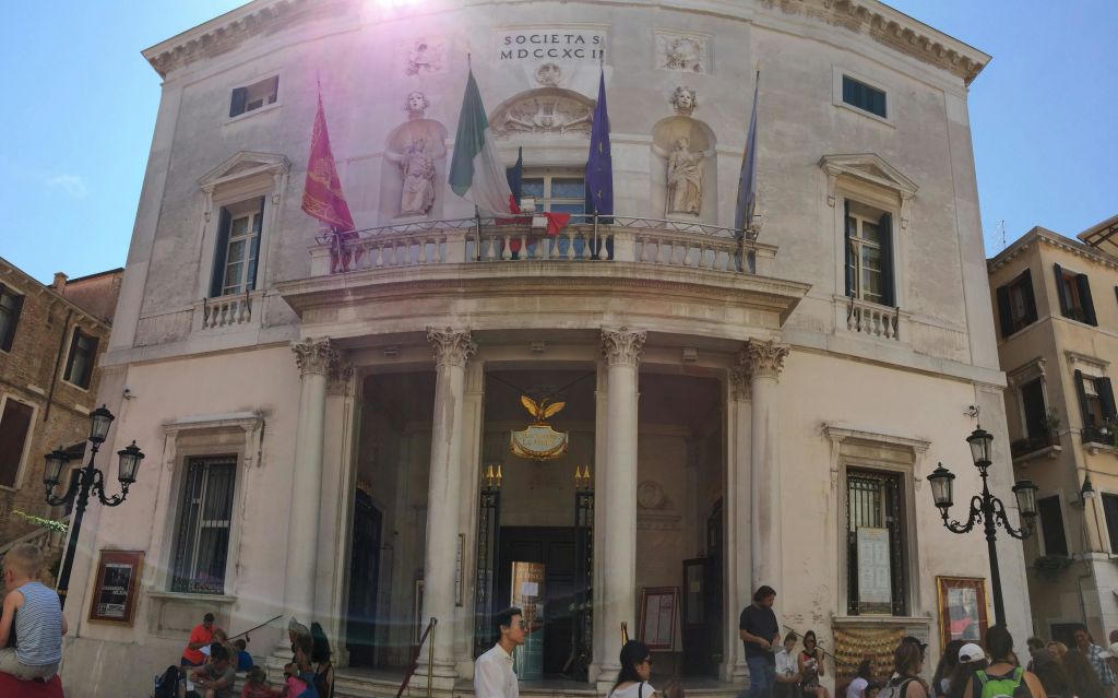 입구 위에 여러 개의 깃발이 장식된 화려한 기둥 모양의 건물 앞에 군중이 모여 있습니다. 햇빛은 렌즈 플레어를 생성합니다.