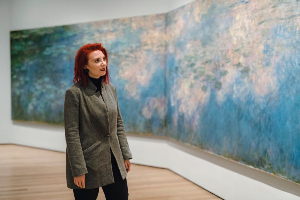 회색 코트를 입은 빨간 머리의 한 여성이 미술관에 서서 벽에 걸린 인상주의 스타일의 커다란 그림을 관찰하고 있습니다.