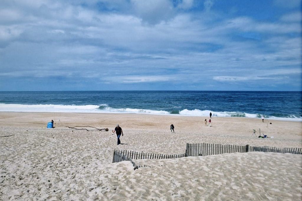 흐린 하늘 아래 바닷가 모래사장을 걷고 있는 사람들로, 앞쪽에는 작은 모래 언덕이 울타리로 둘러싸여 있습니다.