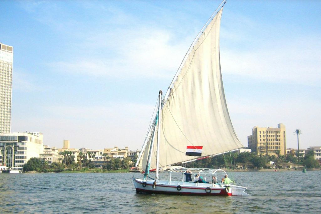 커다란 흰색 돛을 단 보트에 깃발이 달려 있으며, 건물과 나무가 배경으로 보입니다.