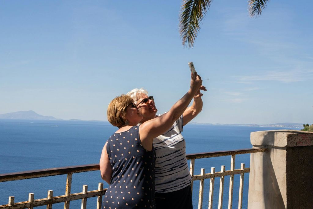 두 사람이 물줄기가 내려다보이는 금속 울타리 옆에 서서 야자수가 보이는 맑은 하늘 아래에서 셀카를 찍고 있습니다.