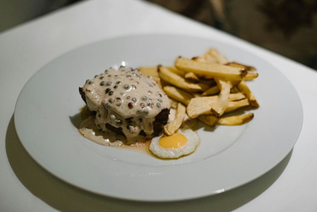 크리미 페퍼콘 소스로 덮인 스테이크를 곁들인 요리로, 흰 접시에 감자튀김과 계란 후라이를 곁들입니다.