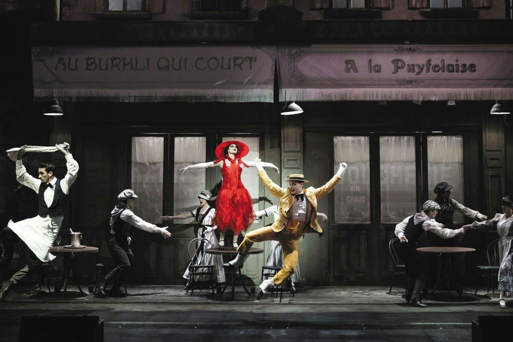 파리의 카페를 닮은 무대에서 생동감 넘치는 의상을 입은 공연자들이 배경으로 춤을 추고 있습니다.