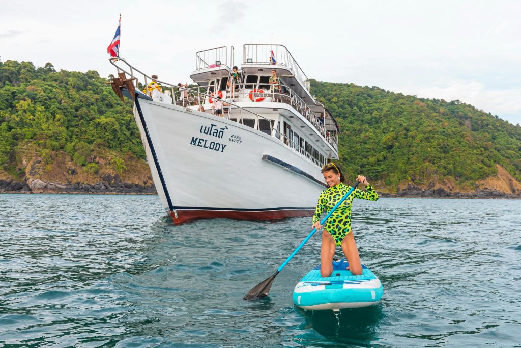 푸른색 스탠드업 패들보드를 타고 '멜로디' 라는 이름의 흰색 보트를 배경으로 한 여성이 노를 젓고 있습니다.