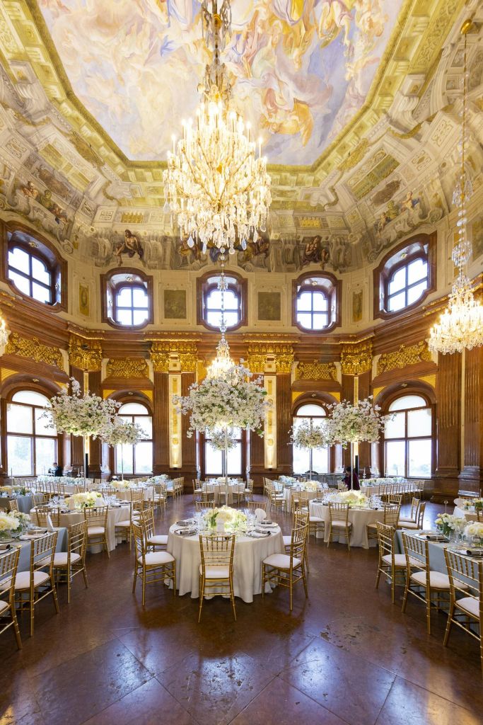 대형 샹들리에, 장식된 천장, 흰색 식탁보가 놓인 원형 테이블, 꽃무늬 센터피스가 있는 호화로운 연회장.