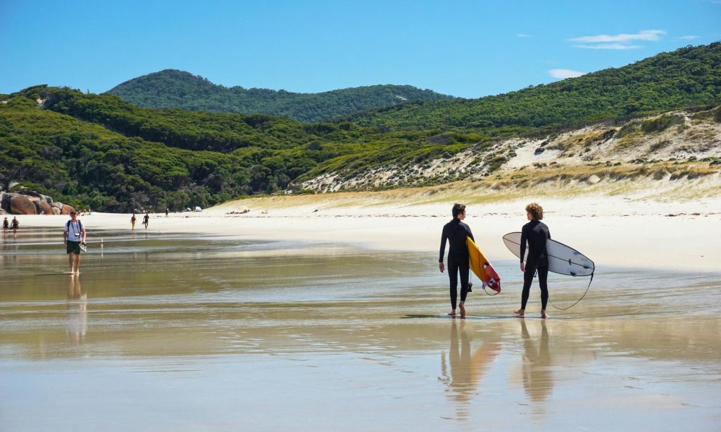 보드를 든 두 명의 서퍼가 모래 해변을 따라 바다를 향해 걸어가고 있습니다. 푸르른 언덕이 배경으로 보입니다.