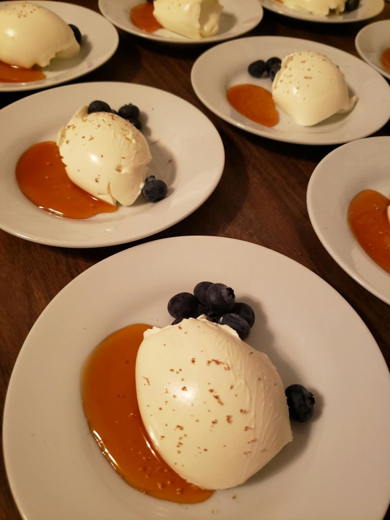 나무 테이블 위에 카라멜 소스와 블루베리가 놓인 판나 코타 한 접시.