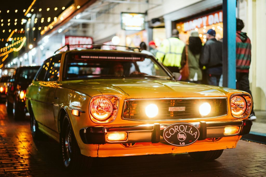 전조등이 켜진 빈티지 옐로우 차량으로, 상점 입구 근처의 줄 조명 아래에 주차되어 있으며 사람들이 바깥에 모여 있습니다.