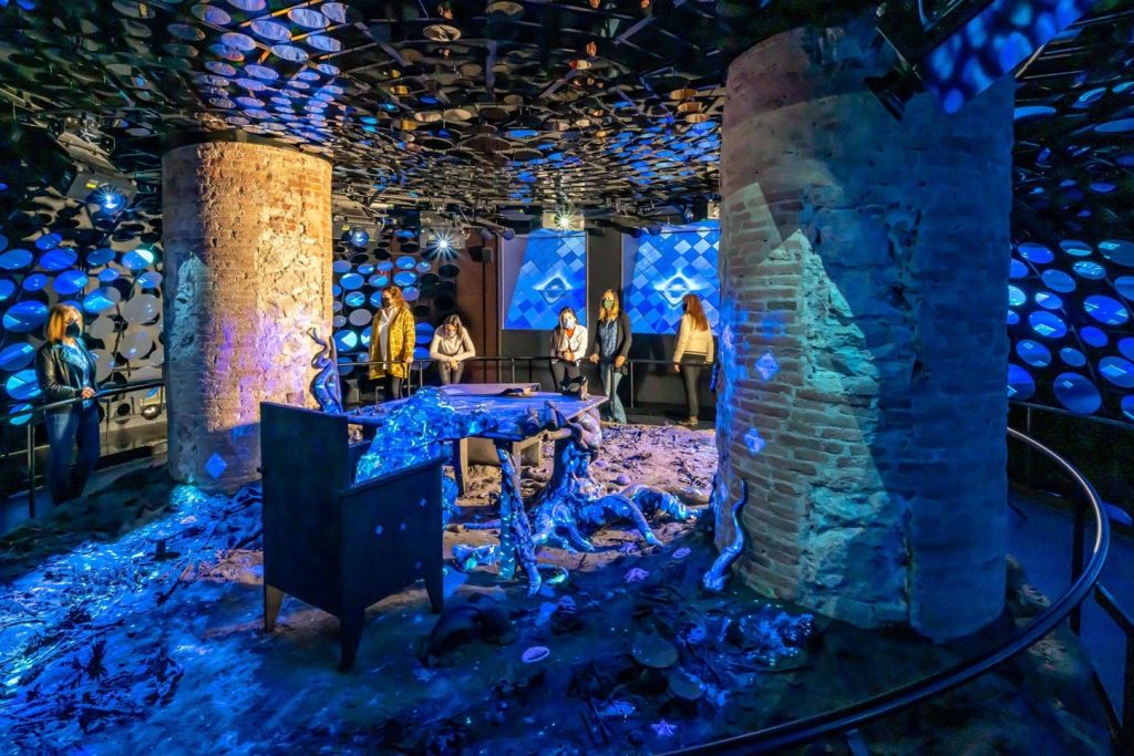 벽돌 기둥과 반사되는 천장 장식이 있는 방에서 사람들은 조명된 추상 예술을 관찰합니다.