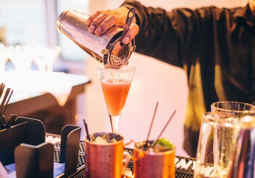 셰이커에서 분홍색 칵테일을 유리잔에 붓고, 뒤쪽에는 구리 머그잔 2개와 바가 있습니다.