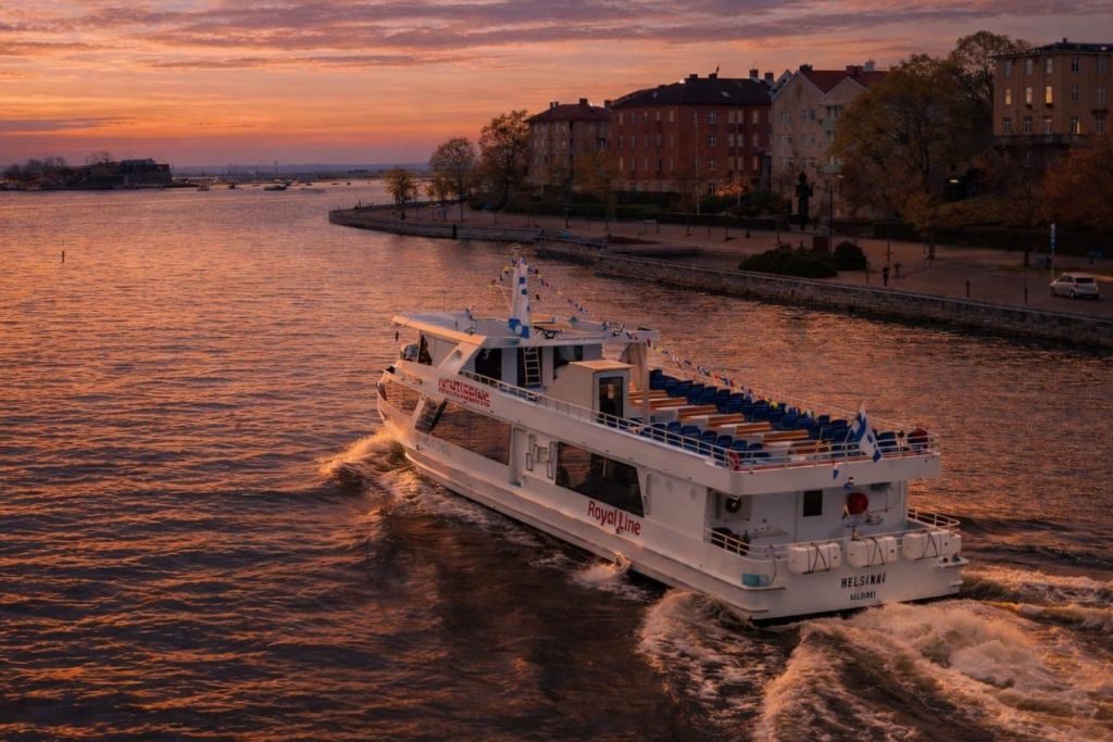 "로얄 라인" 이라고 표시된 흰색 페리가 해안가를 따라 보이는 건물과 나무가 있는 석양의 잔잔한 수로를 항해합니다.