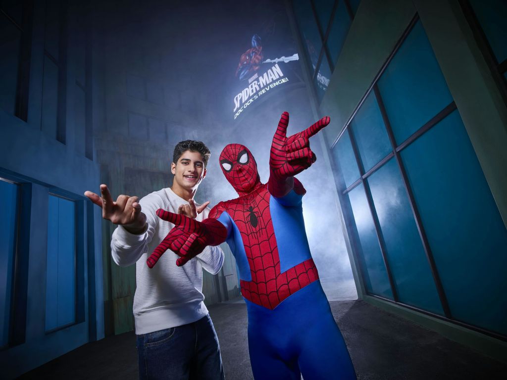 흰색 스웨터를 입은 사람이 스파이더맨 표지판을 배경으로 도시 풍경에서 스파이더맨 의상을 입은 사람과 포즈를 취하고 있습니다.