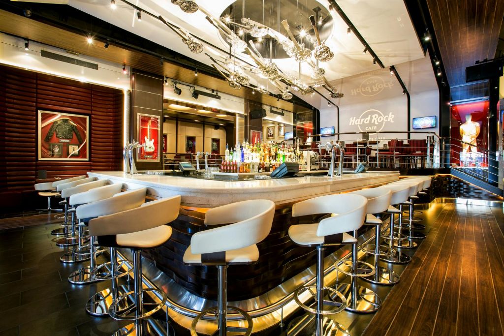 벽에 흰색 스툴, 중앙 집중식 병 디스플레이, 현대적인 장식, "Hard Rock Café" 로고가 있는 바입니다.
