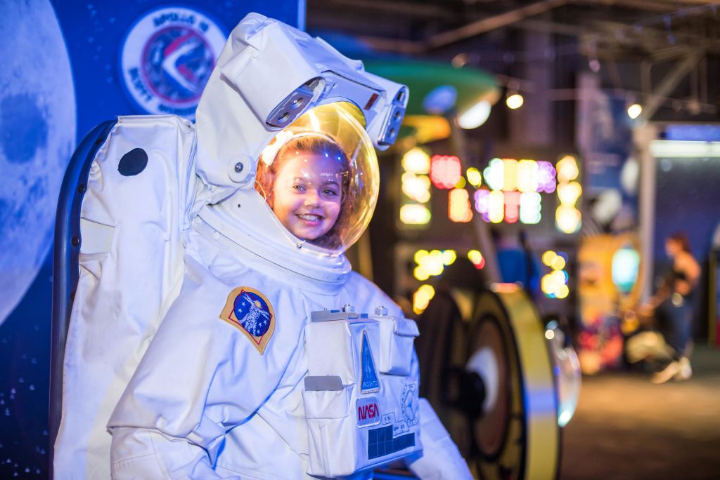 다채로운 조명을 배경으로 우주를 주제로 한 전시관에서 커다란 우주비행사복을 입고 미소를 짓고 있는 어린이.