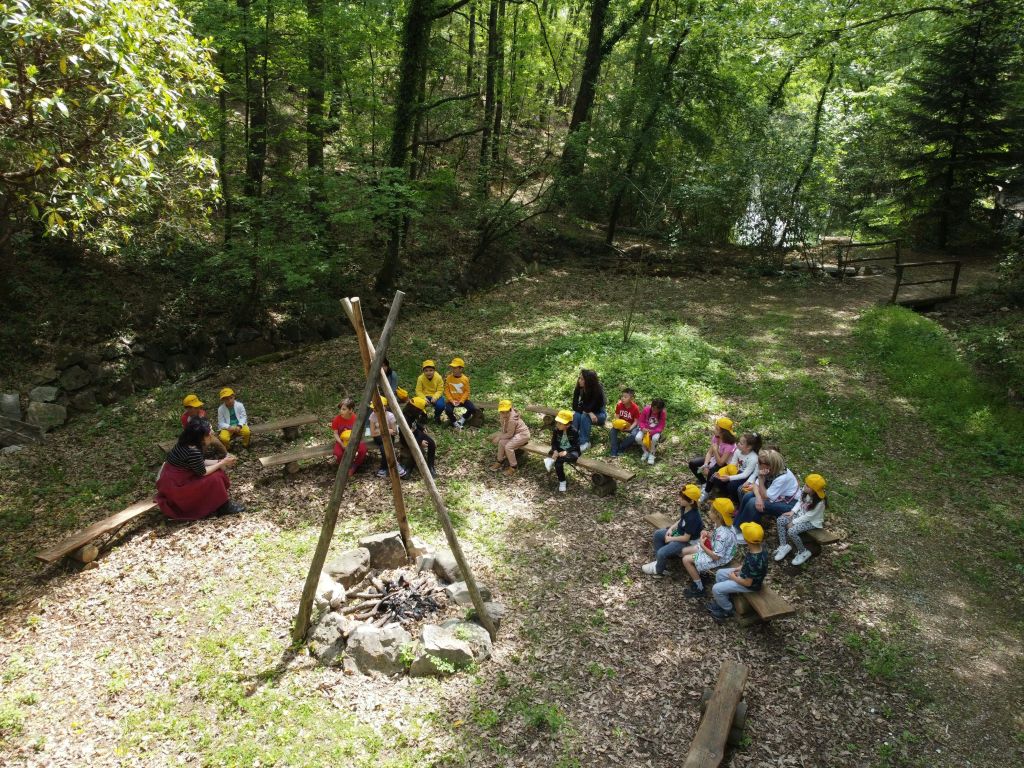 임시 캠프파이어 시설 근처에서 두 명의 어른의 말을 주의 깊게 들으며 숲속의 벤치에 앉아 있는 어린이들.