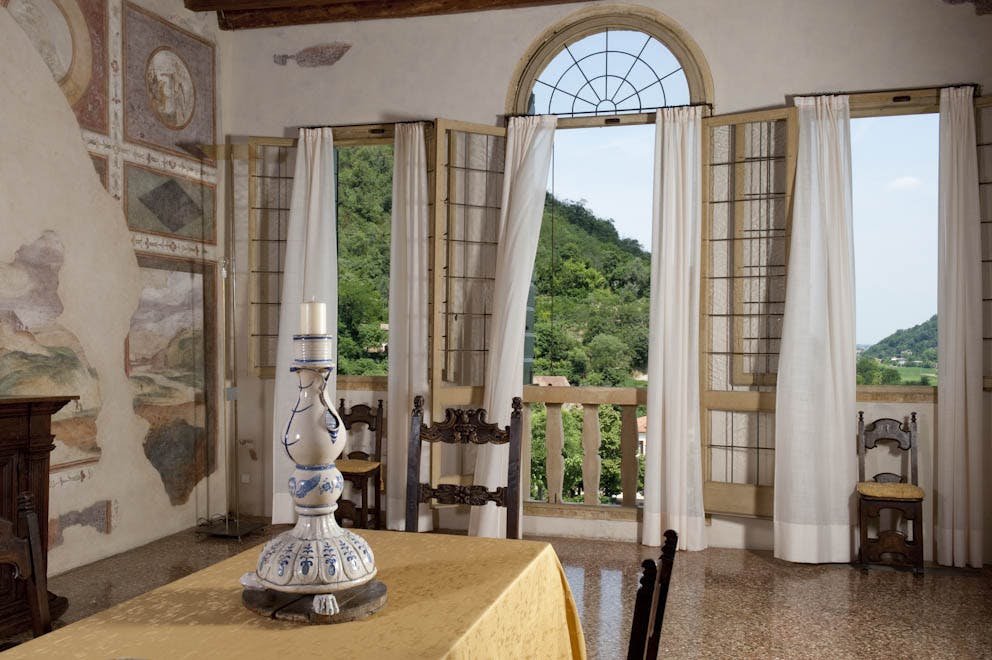 커다란 아치형 창문, 오픈 셔터, 캔들 홀더가 있는 테이블, 바깥의 푸르른 풍경이 보이는 객실입니다.