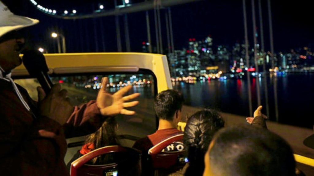 도시의 스카이라인과 다리를 배경으로 한 야간 오픈탑 버스를 탄 사람들.