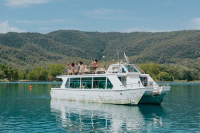 푸른 숲이 우거진 언덕을 배경으로 고요한 호수에서 상부 갑판에 사람들이 타고 있는 흰색 보트를 타고 항해하고 있습니다.