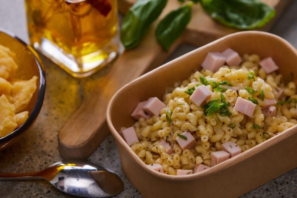 햄 큐브와 허브, 숟가락, 음료, 바질이 들어간 마카로니 샐러드 한 그릇이 나무 판자에 담겨 있습니다.