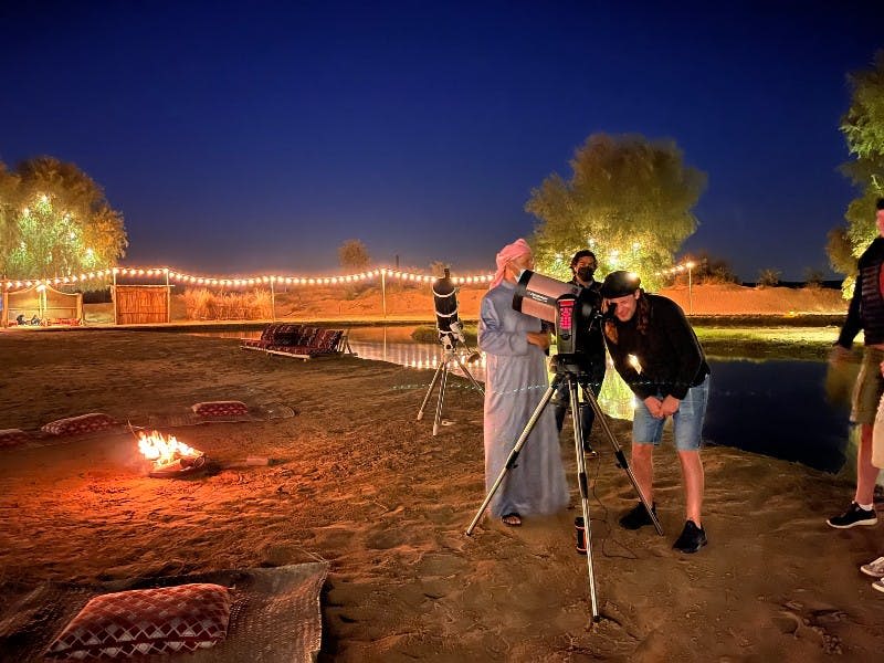 두 사람이 밤에 작은 캠프파이어 근처에서 망원경을 들여다보고 있으며, 줄 조명과 나무로 둘러싸여 있습니다.