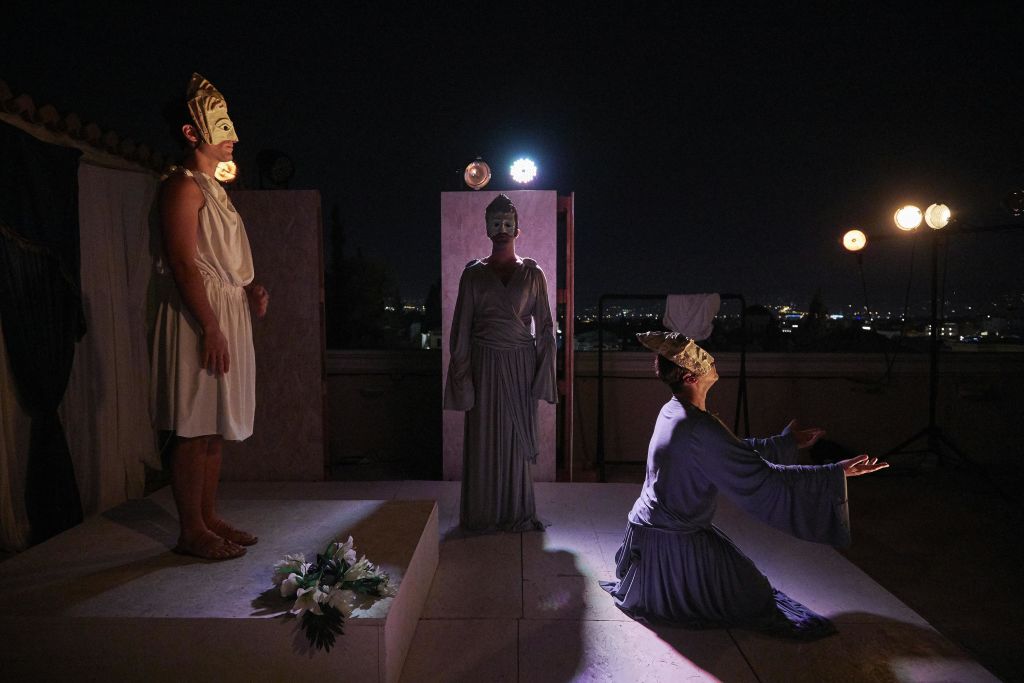 마스크를 쓴 세 명의 배우가 밤에 무대에서 극적인 장면을 연출합니다. 한 명은 무릎을 꿇고 두 명은 서 있습니다. 소품에는 꽃과 빨래판이 포함됩니다.