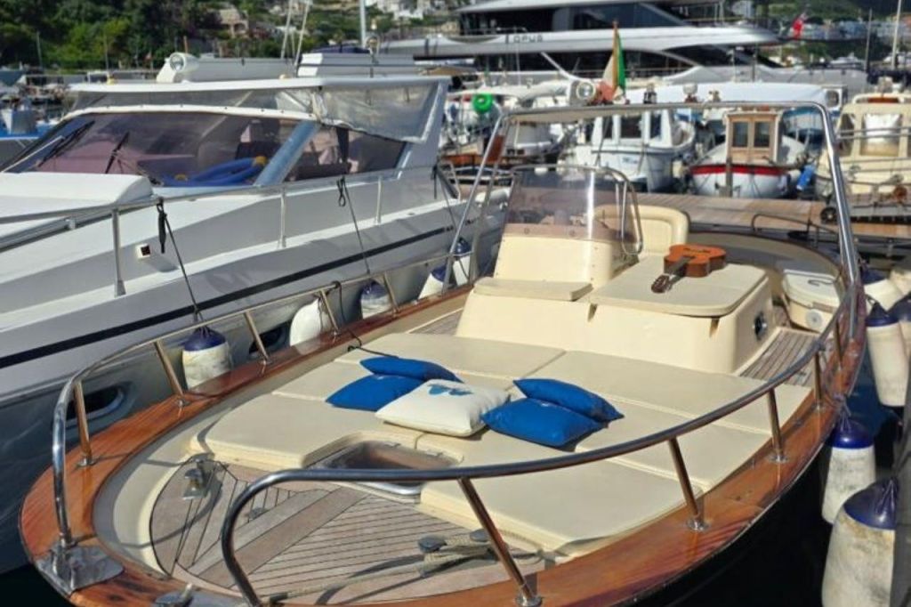베이지색 데크 쿠션, 파란색과 흰색 베개, 좌석 공간에 기타가 있는 도킹 요트입니다. 배경에 다른 보트.