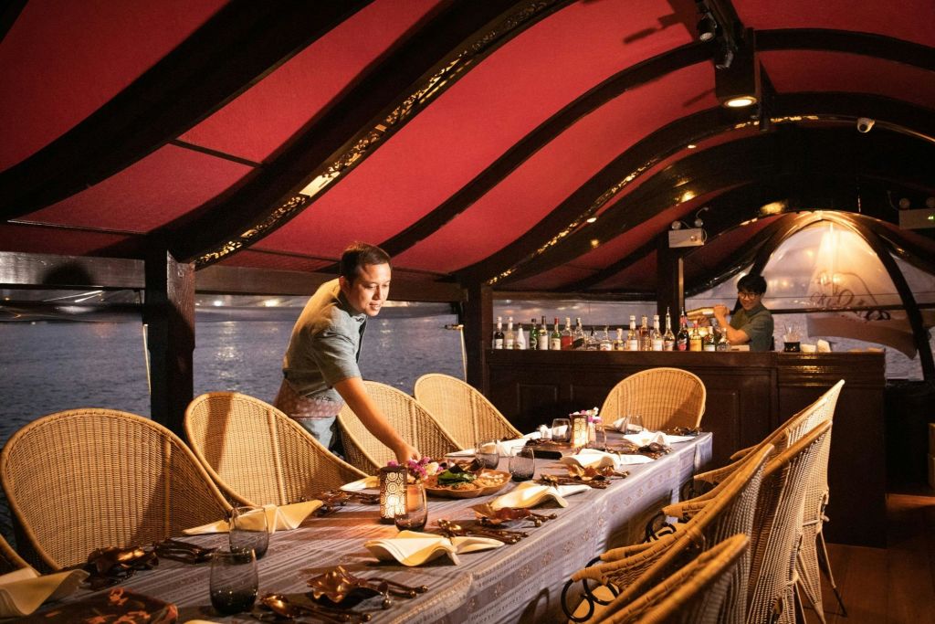 한 사람이 보트의 빨간색 캐노피 아래에 화려한 장식이 놓인 테이블을 배경으로 바와 물이 보입니다.