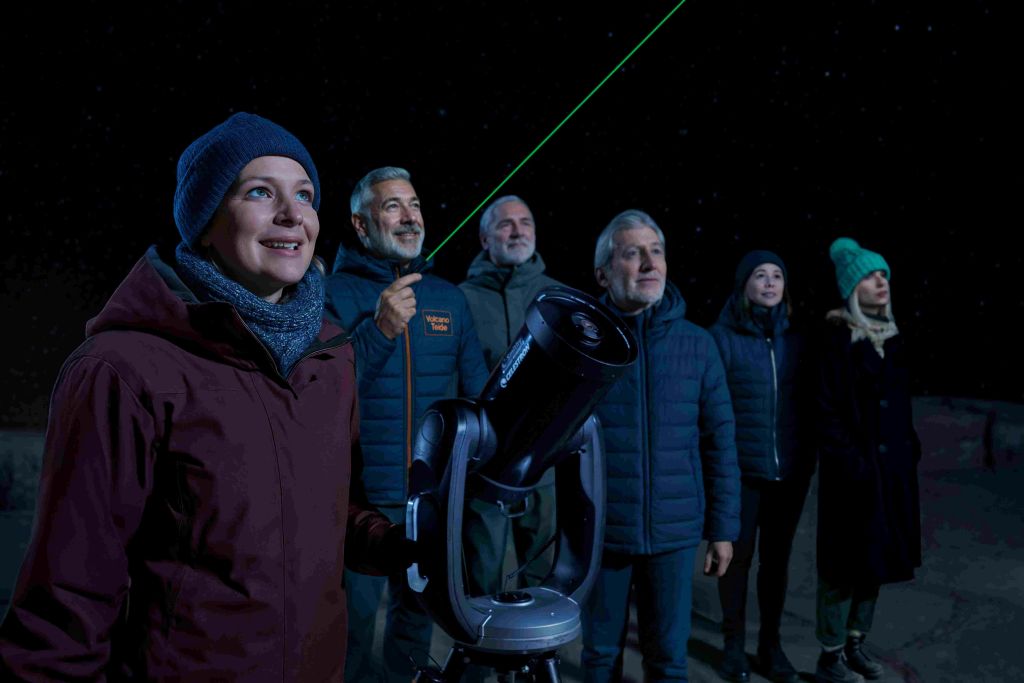 빨간 재킷을 입은 여성이 망원경을 들고 미소를 짓고 있습니다. 재킷을 입은 세 남자가 그녀의 뒤에 서서 한 남자가 밤하늘을 향해 초록색 레이저를 가리키고 있습니다.