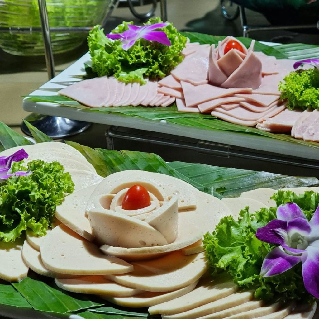 바나나 잎에 양상추, 보라색 꽃, 체리 토마토를 장식한 얇게 썬 델리 고기 두 접시.