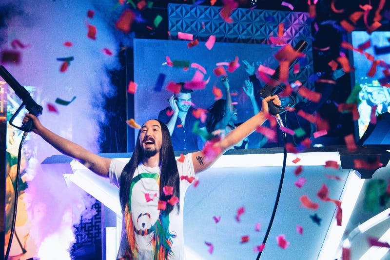 화려한 색종이 무대가 펼쳐진 무대에서 두 팔을 쭉 뻗은 채 DJ 부스를 배경으로 축하무대가 펼쳐집니다.