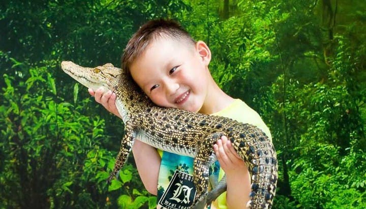 웃는 아이가 울창한 녹색 정글 배경 앞에 서서 작은 악어를 뺨에 붙잡고 있습니다.