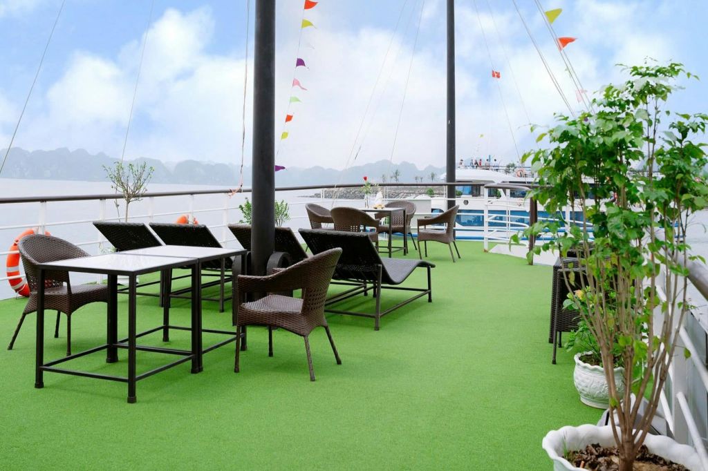 고리 버들 의자, 테이블, 화분, 화려한 깃발이 있는 녹색 바닥의 야외 데크. 보트와 산을 배경으로 진행됩니다.