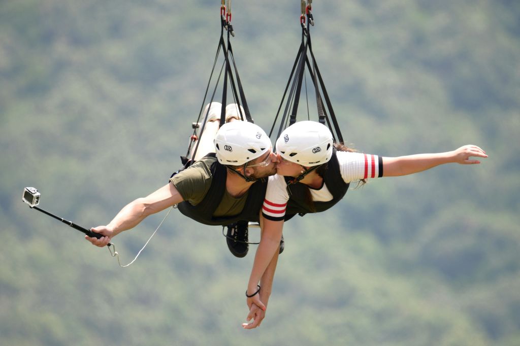 푸르스름한 녹색 풍경을 배경으로 집라인 하네스에 매달린 채 헬멧을 쓴 부부가 키스하고 있습니다.