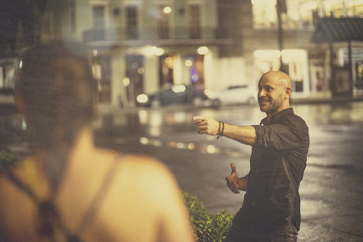 검은 셔츠를 입은 한 남성이 흐릿한 건물과 차를 배경으로 젖은 거리에서 미소를 짓고 있습니다.