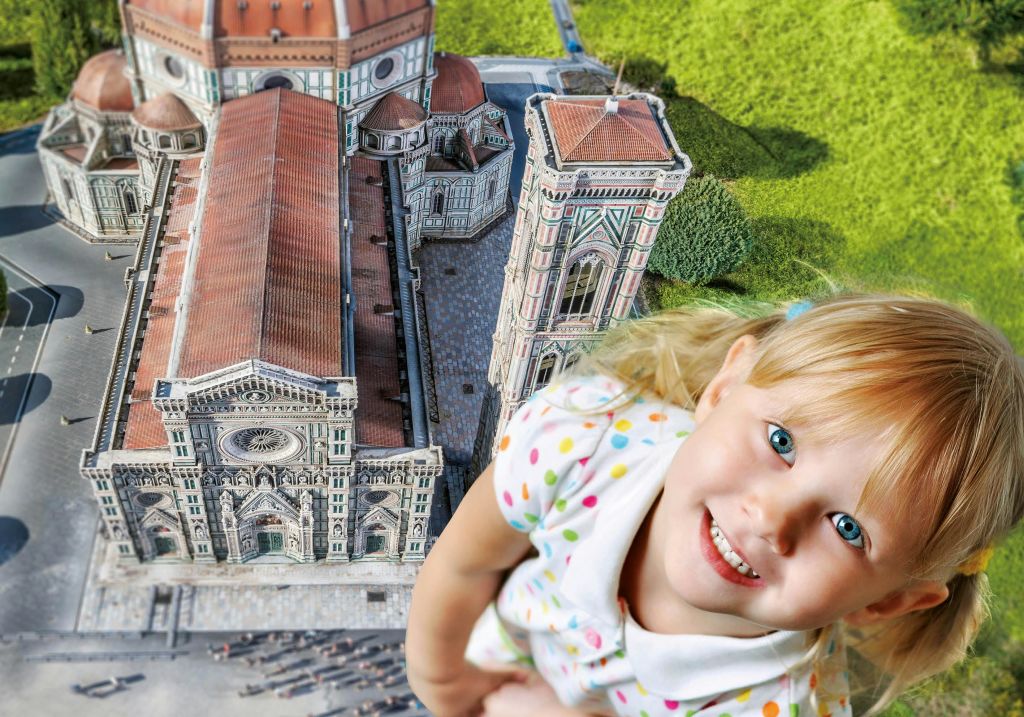 역사적인 건물과 울창한 녹지의 상세한 미니어처 모델 옆에 금발의 미소 짓는 어린 소녀가 거대한 모습으로 나타납니다.