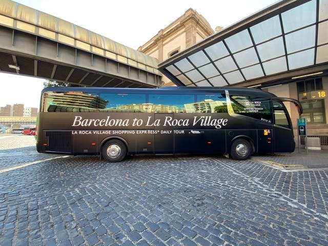 현대식 건물과 고가도로 근처의 자갈길에 '바르셀로나 ~ 라 로카 빌리지' 라는 이름의 검은색 투어 버스가 주차되어 있습니다.