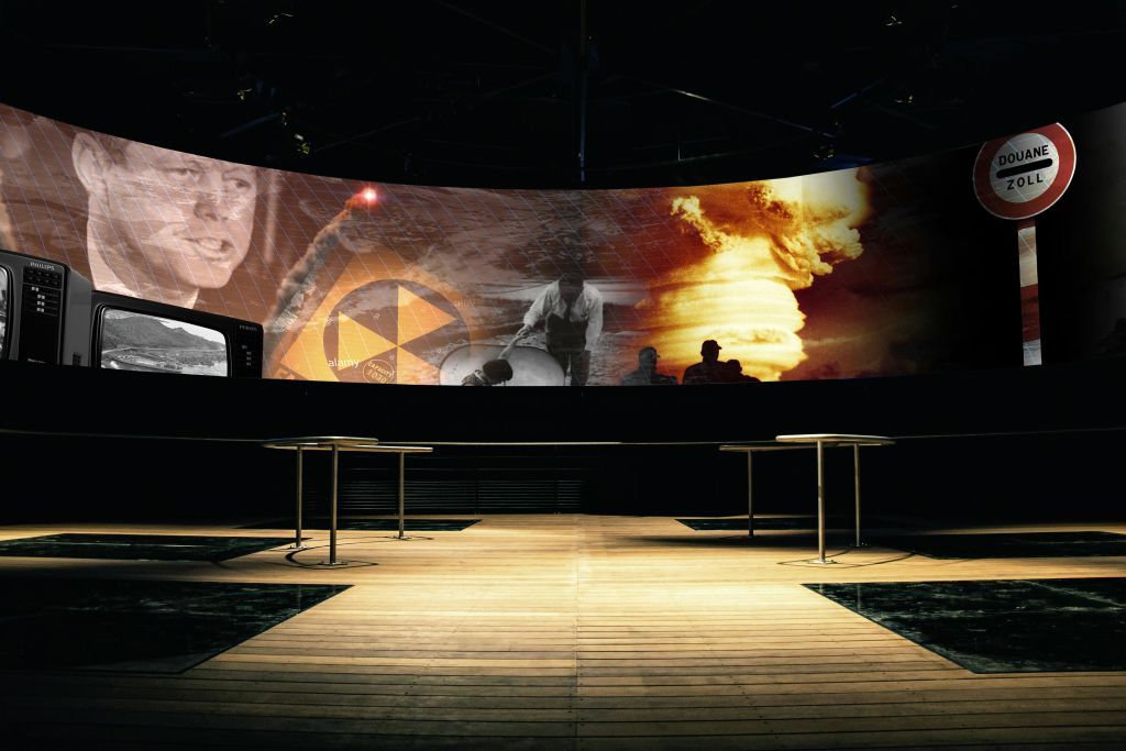 나무 바닥과 빈 테이블이 있는 방에 사람, 핵 기호, 미사일 등 다양한 이미지가 표시된 곡선형 화면.